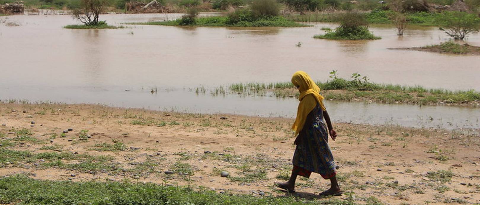 Young Sudanese girl walking along shallow river with dried shrubs and trees in the horizon.
