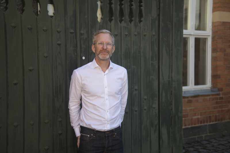 Portrait picture of Henrik Österblom, wearing a white shirt, standing in front of a green door