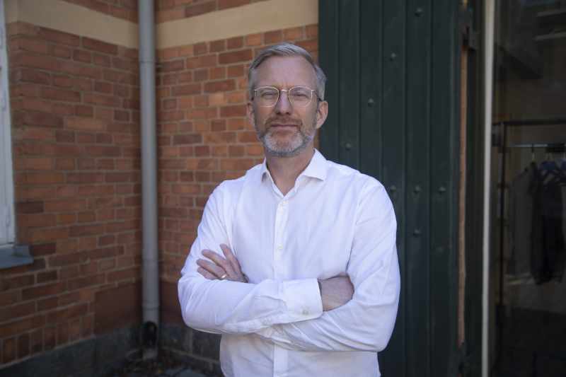 Portrait image of Henrik Österblom, wearing a white shirt standing in front of a red brick wall